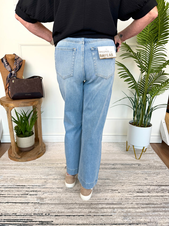 A person shown from the back wearing light-wash blue jeans with a visible back patch that reads BAVEAS, standing indoors on a patterned rug with plants and furniture in the background.