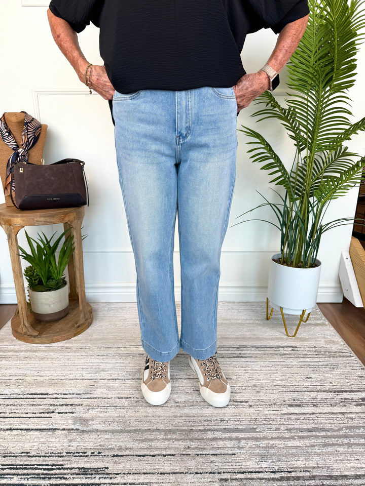 A person wearing light-wash blue straight-leg jeans, a black top, and beige sneakers, standing indoors on a gray patterned rug with a potted palm and a small wooden side table in the background.
