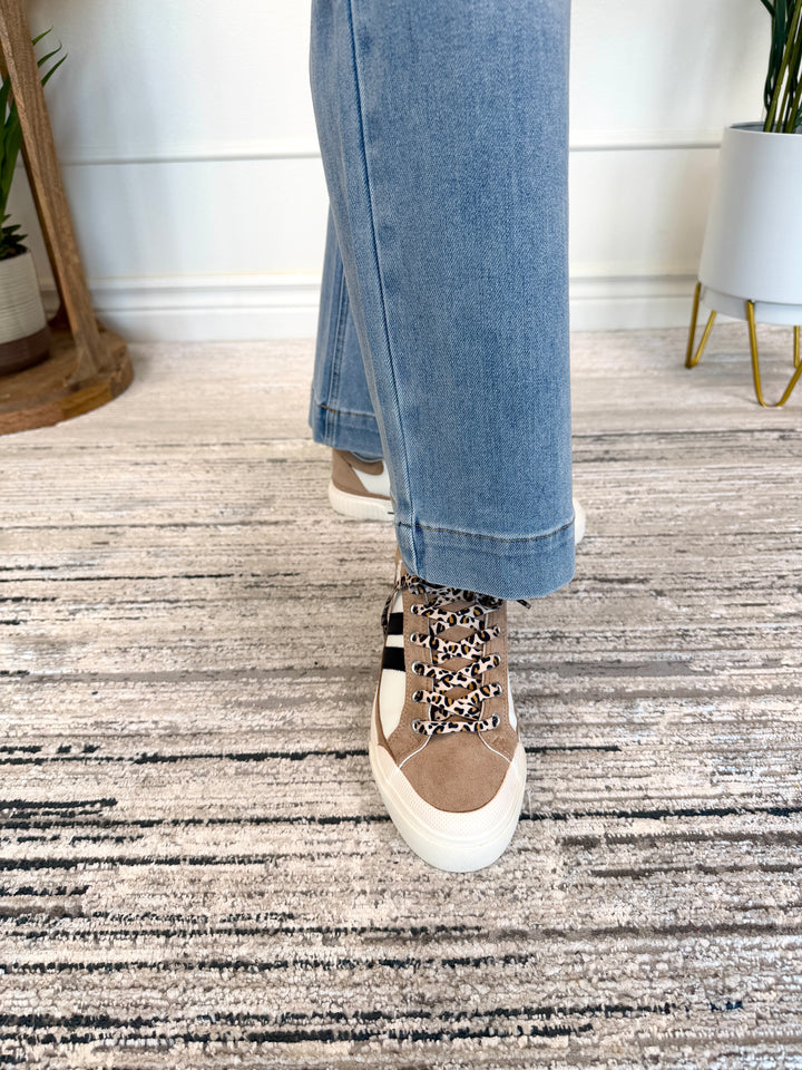 A person wearing light blue jeans and a tan low-top sneaker with leopard-print laces, standing on a textured gray rug in an indoor setting.
