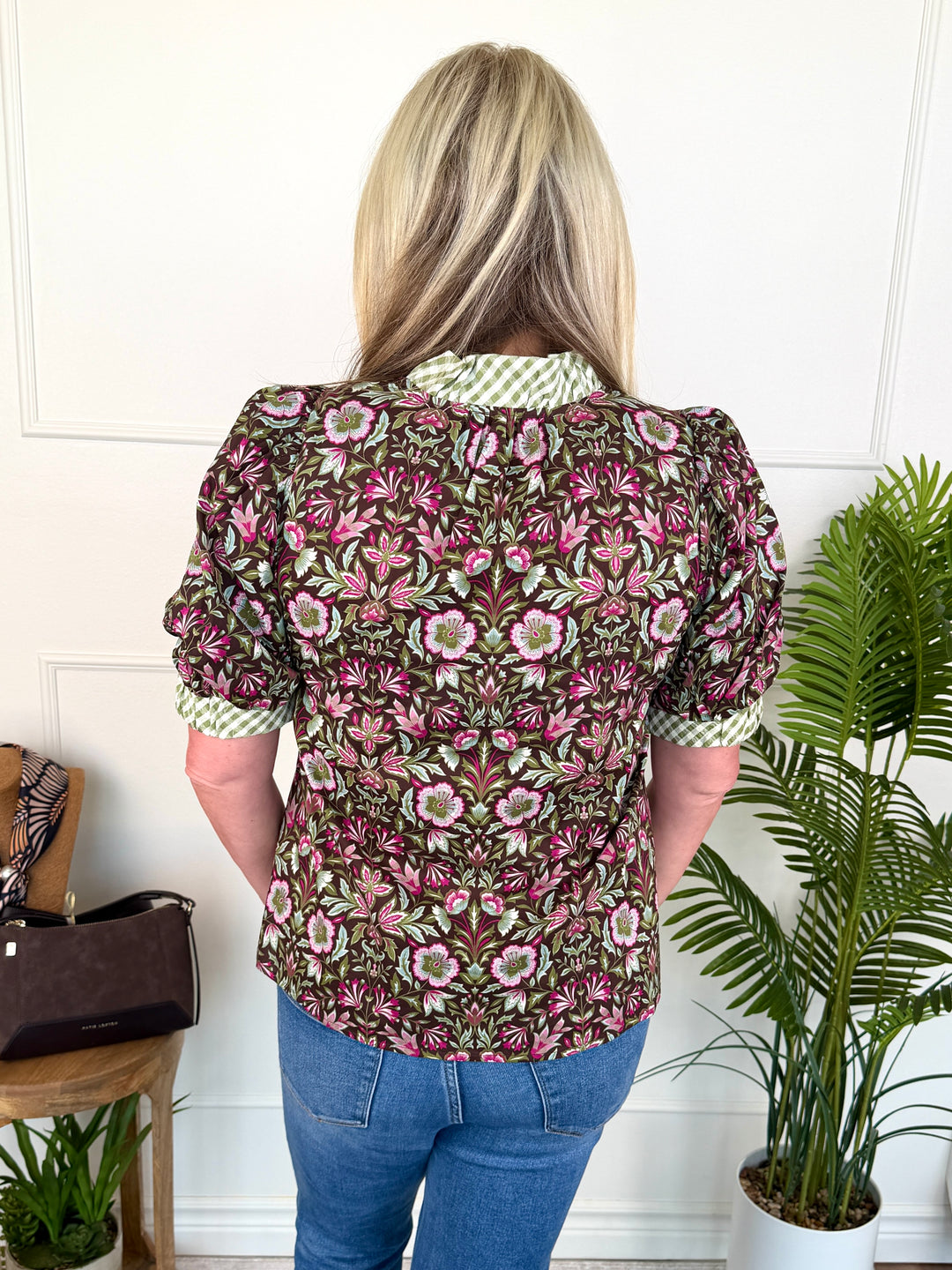 A woman viewed from behind wearing a short-sleeve floral blouse with puff sleeves and a green-and-white check collar, paired with blue jeans, standing indoors against a white paneled wall with potted plants nearby.