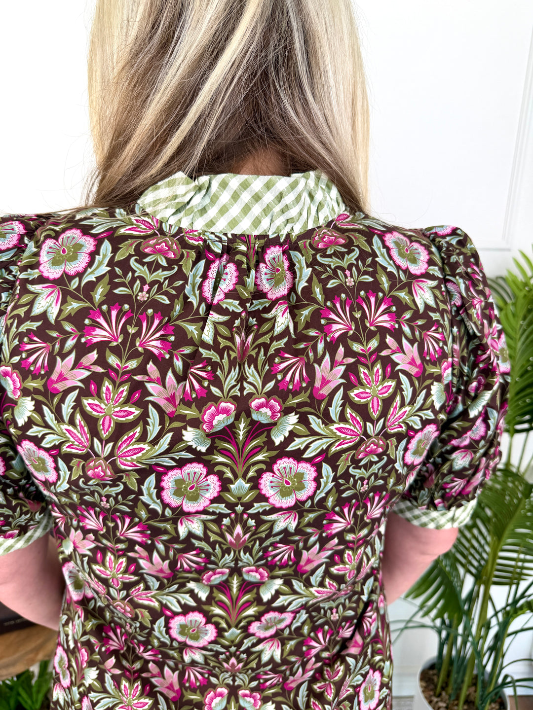 A woman viewed from behind wearing a short-sleeve puff-sleeve blouse with a dense floral pattern in olive green, pink, and white, featuring a contrasting green and white gingham collar.