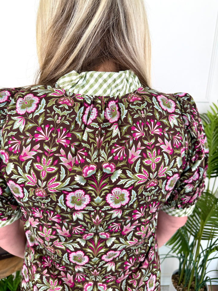 A woman viewed from behind wearing a short-sleeve puff-sleeve blouse with a dense floral pattern in olive green, pink, and white, featuring a contrasting green and white gingham collar.