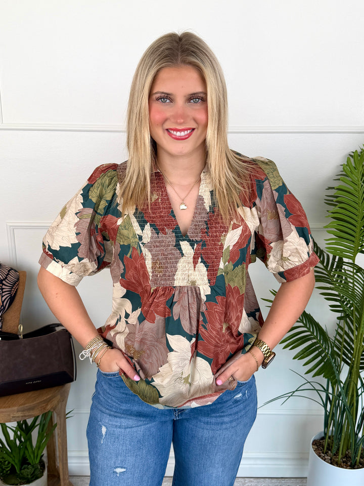A smiling blonde woman wearing a short-sleeve, smocked-neck blouse with a brown, beige, and green botanical leaf pattern, paired with blue jeans, standing in front of a white paneled wall with indoor plants.