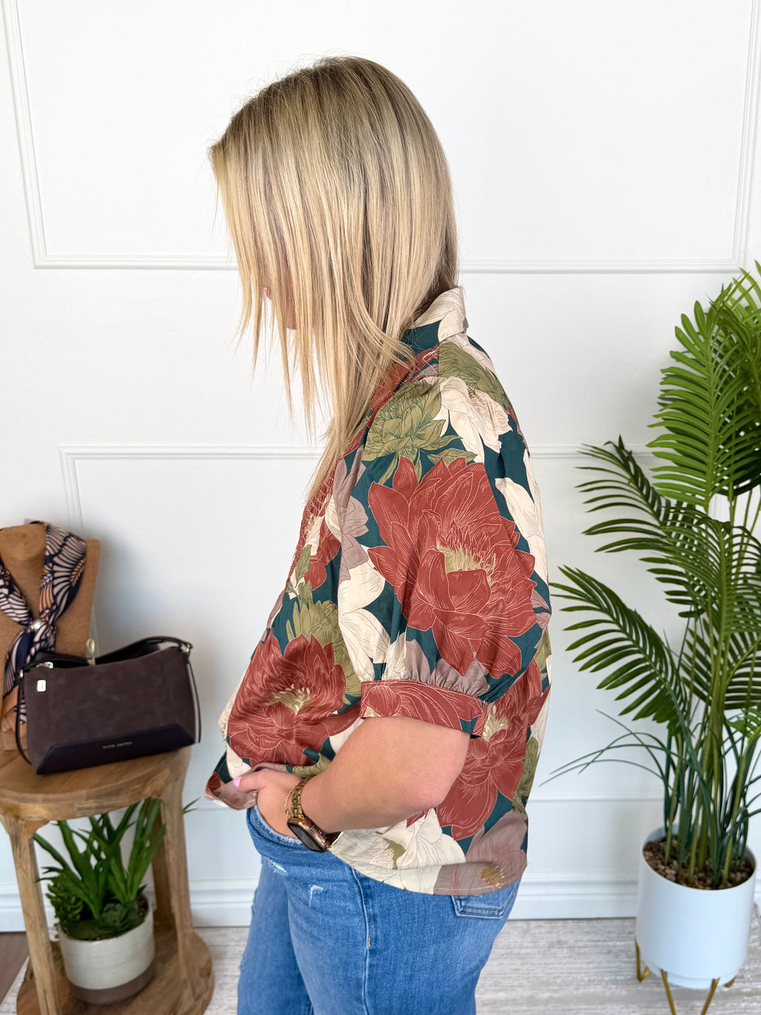 A woman shown from the back wearing a short-sleeve button-up shirt with a large floral print in rust red, beige, and green, paired with blue jeans in a bright indoor setting.