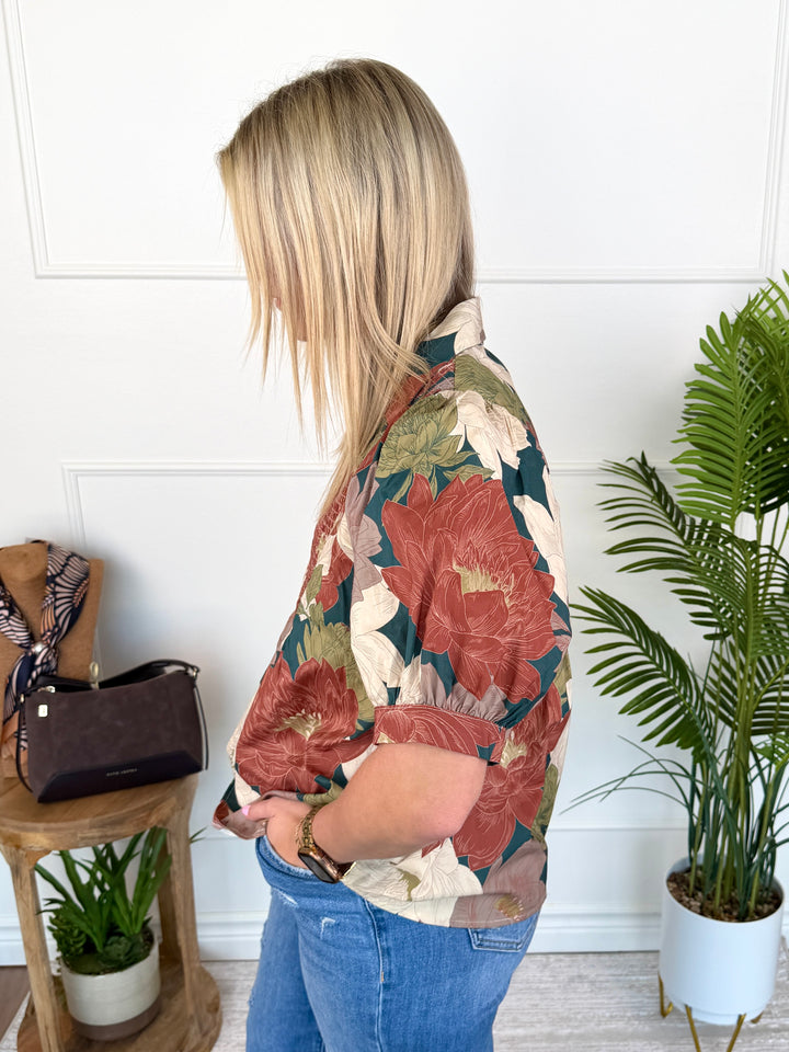 A woman shown from the back wearing a short-sleeve button-up shirt with a large floral print in rust red, beige, and green, paired with blue jeans in a bright indoor setting.