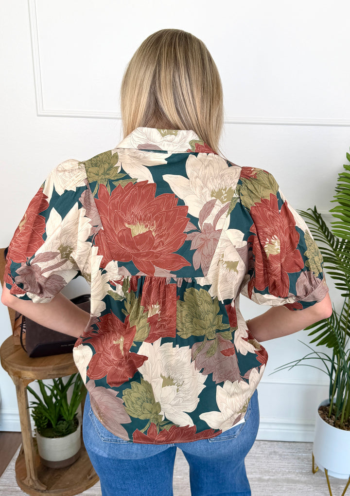 A woman viewed from behind wearing a short-sleeve button-up blouse with a large floral print in muted red, cream, olive green, and dark teal, paired with blue jeans in an indoor setting.