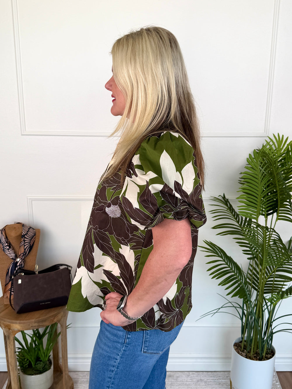 A woman with blonde hair is shown from the side, wearing a short-sleeve button-up blouse with a green, brown, and cream floral pattern, paired with blue jeans. She is standing in a room with a white paneled wall, next to a wooden side table with a handbag and a potted plant.