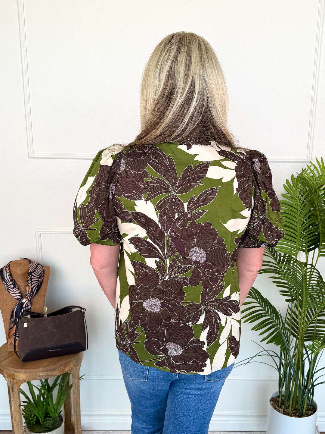 A woman viewed from behind wearing a short-sleeve blouse with a bold floral pattern in olive green, dark brown, and cream. She is standing in a bright room with a white paneled wall, next to a wooden stool with a handbag and a potted palm plant.