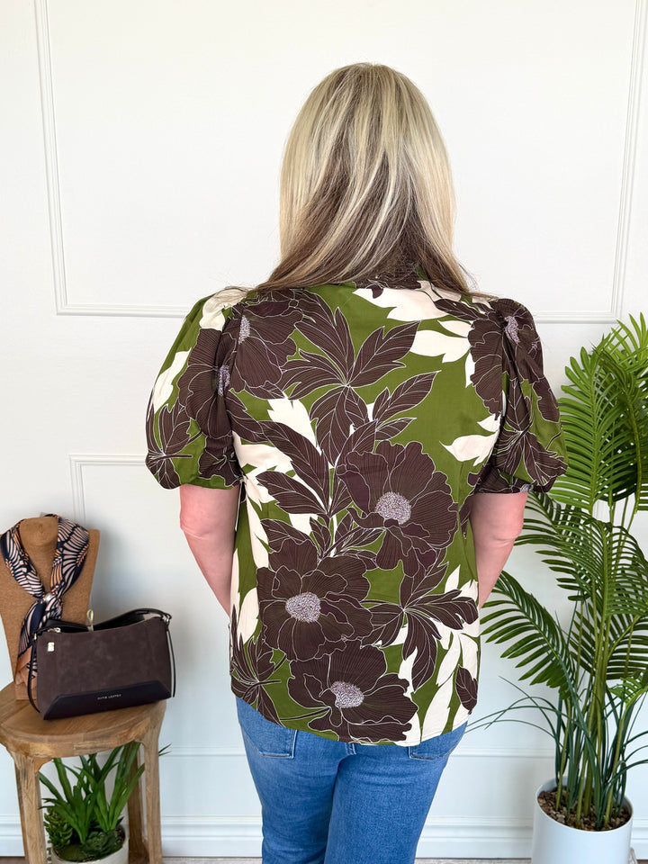 A woman viewed from behind wearing a short-sleeve blouse with a bold floral pattern in olive green, dark brown, and cream. She is standing in a bright room with a white paneled wall, next to a wooden stool with a handbag and a potted palm plant.