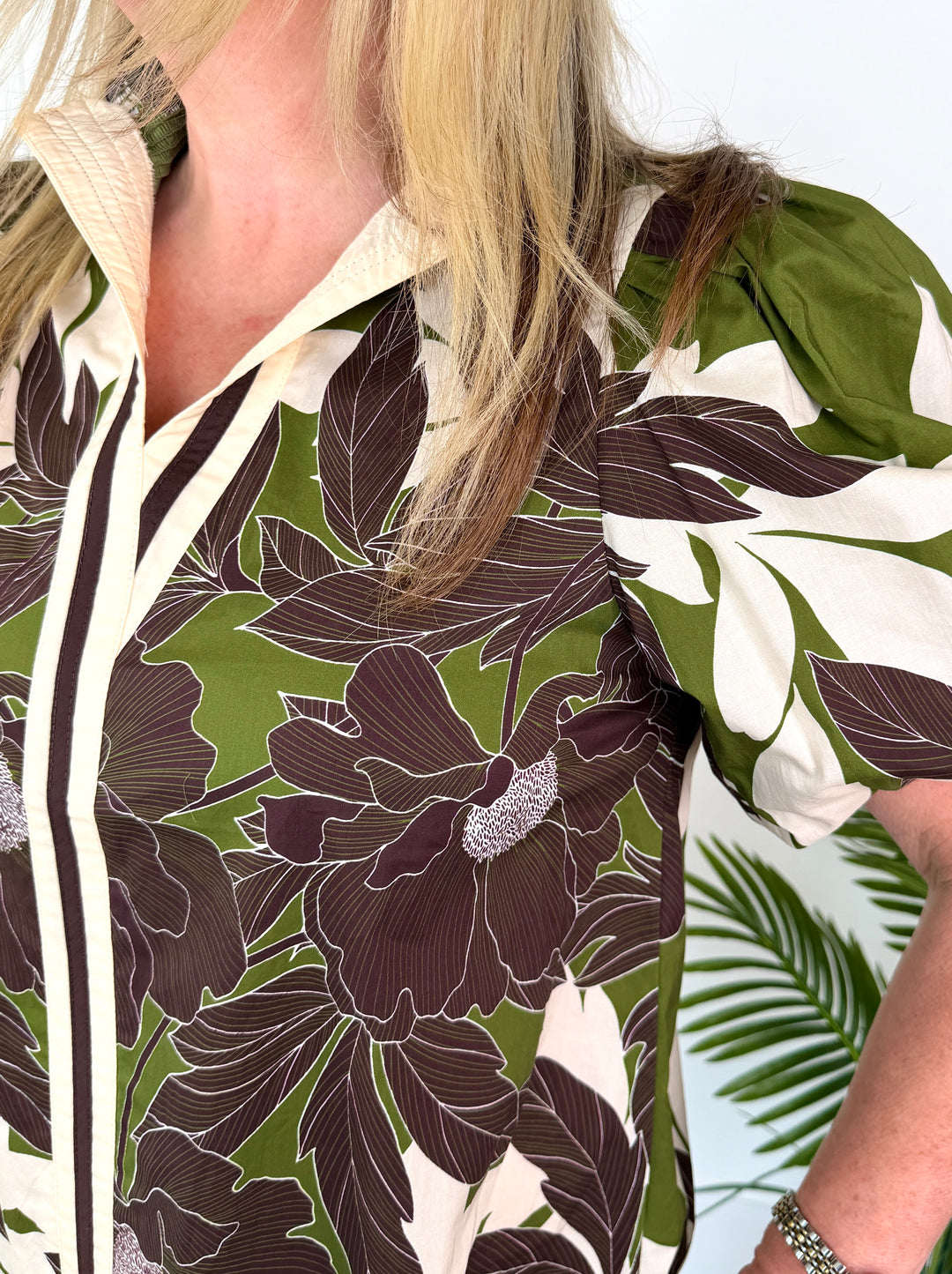 A close-up of a woman wearing a short-sleeve button-front blouse with a bold tropical floral print in olive green, cream, and dark brown.