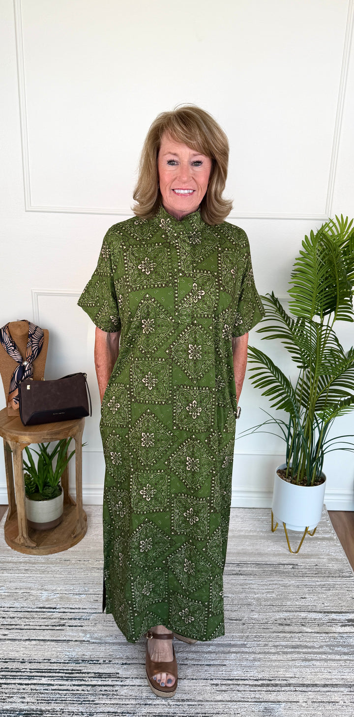 A woman modeling a short-sleeved, high-neck maxi dress in olive green with a small floral pattern, standing in a brightly lit room with potted plants and a wooden side table.