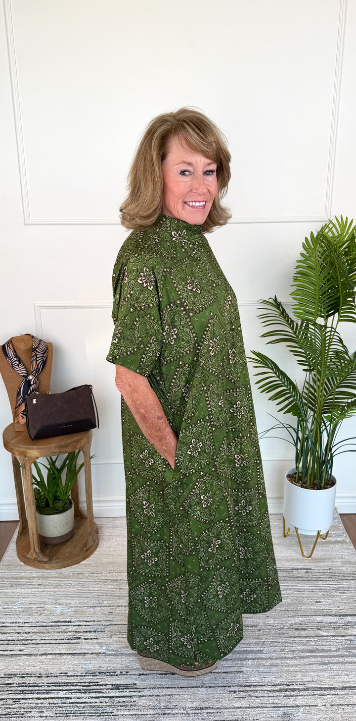 A woman modeling a short-sleeve, high-neck, loose-fitting maxi dress with a green and beige floral pattern, standing in a styled indoor setting with a wooden side table and a potted palm.
