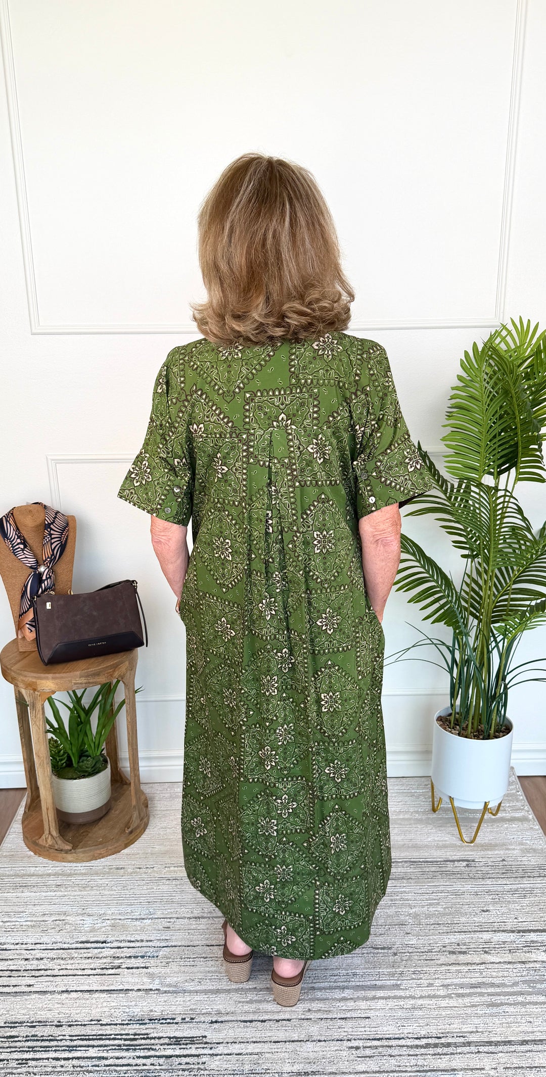 A woman viewed from behind wearing a short-sleeve olive green midi dress with a small floral print, standing in a room with a white paneled wall, a potted plant, and a small wooden side table.