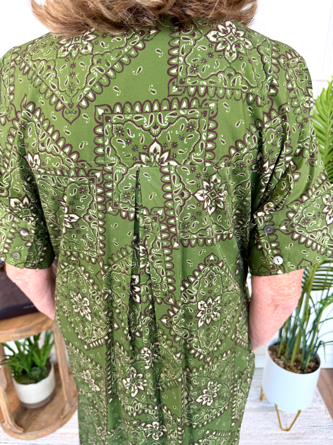 A person wearing a short-sleeve green top with an intricate brown and beige paisley and floral pattern, shown from the back in an indoor setting with potted plants.