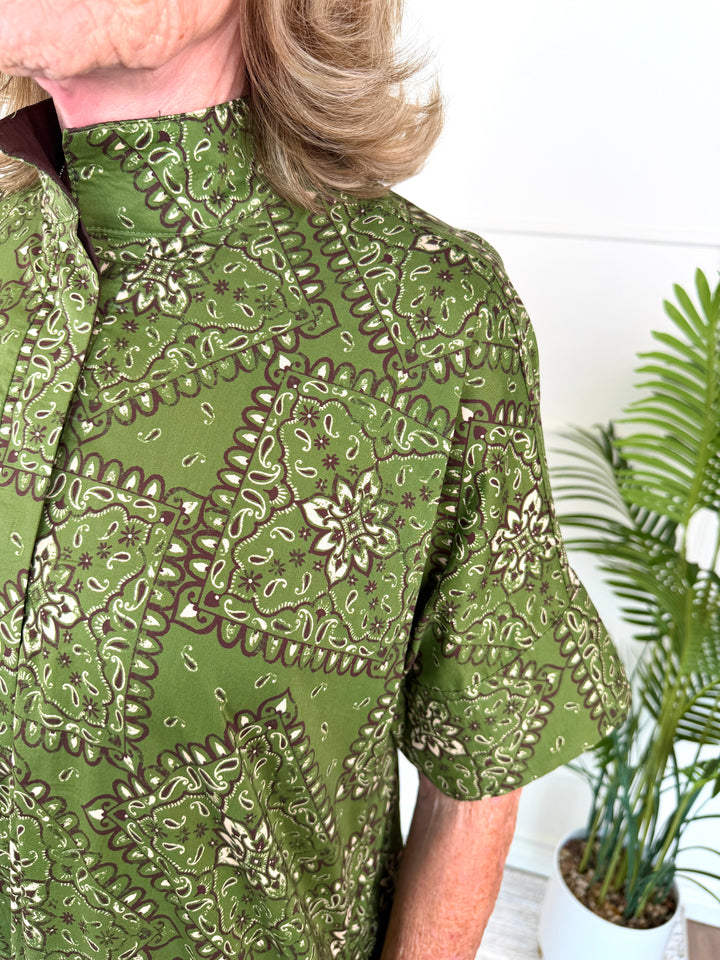A close-up of a person wearing a short-sleeve button-front shirt with an olive green and brown paisley pattern, standing indoors next to a potted plant.