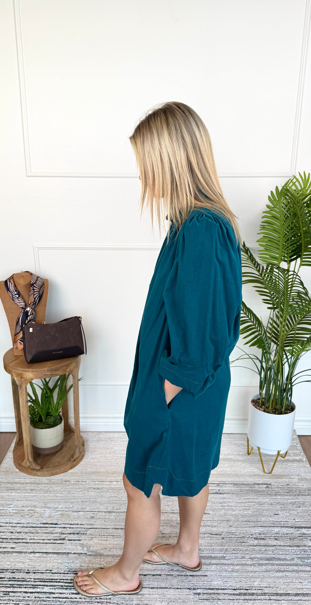 A woman wearing a teal blue-green long-sleeve tunic dress with side pockets, standing in a bright room with a white paneled wall, a potted plant, and a small wooden side table.