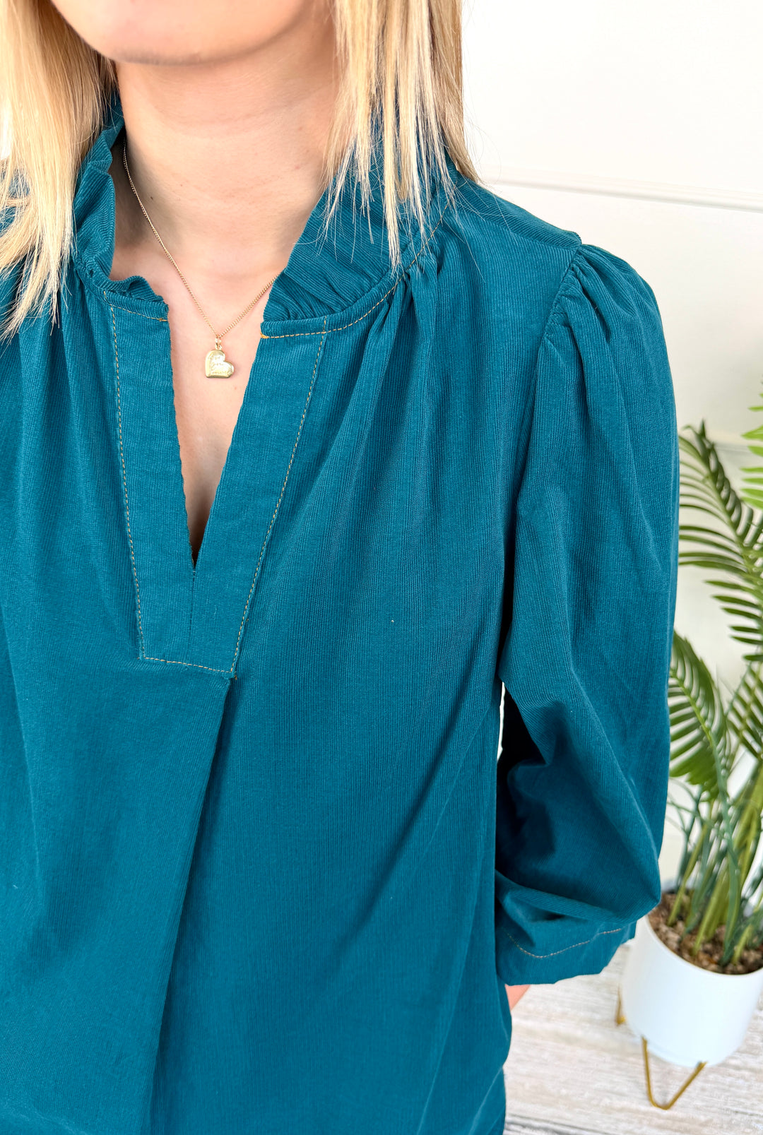 A close-up of a woman wearing a teal blue long-sleeve blouse with a gathered high neck and puff sleeves, paired with a delicate gold necklace.