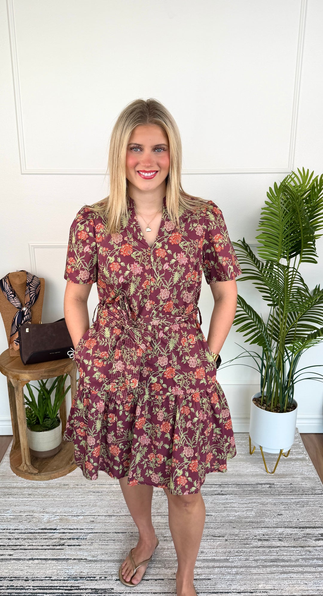 A woman modeling a short-sleeve floral dress with a ruffled tiered hem, standing in a bright room with a white paneled wall, a potted plant, and a small side table.