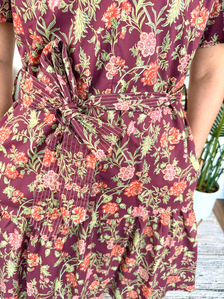 Close-up of a floral dress with a tie waist belt, featuring a burgundy base with pink, orange, and green botanical print.