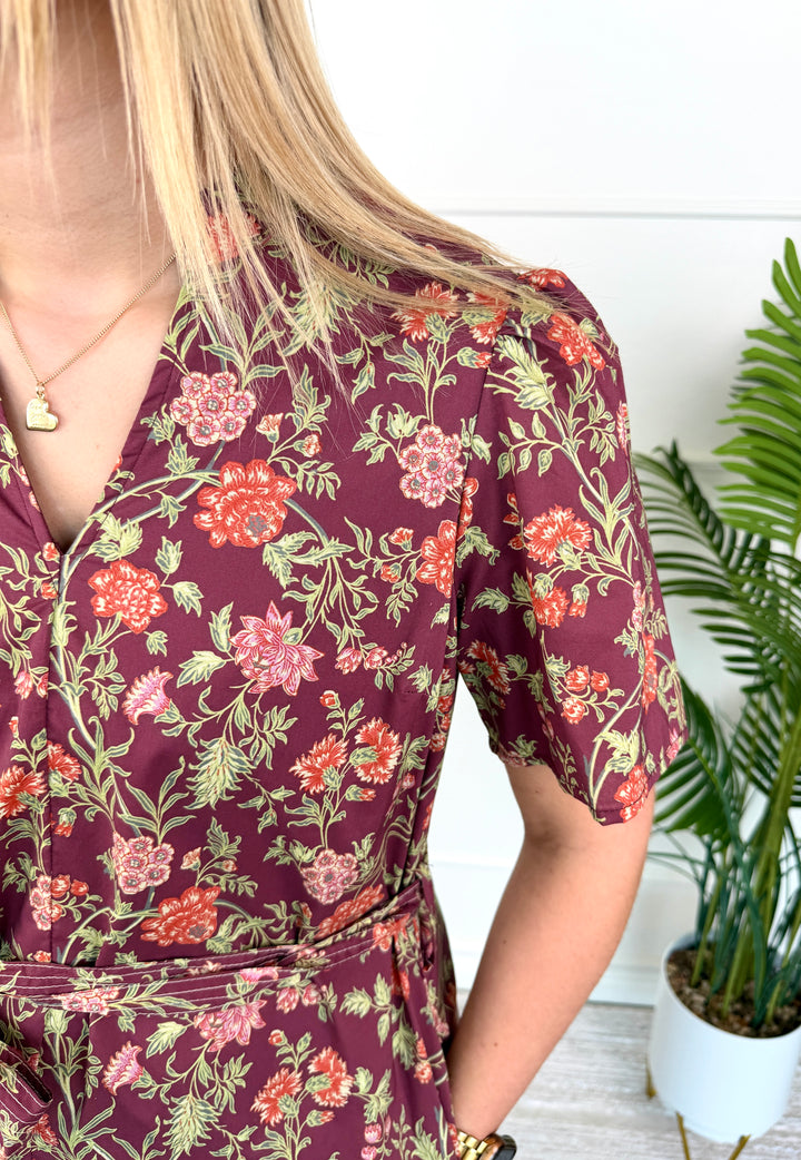 A close-up of a woman wearing a short-sleeve wrap dress with a burgundy base and a vibrant floral pattern of pink, orange, and green flowers, standing next to a potted plant.
