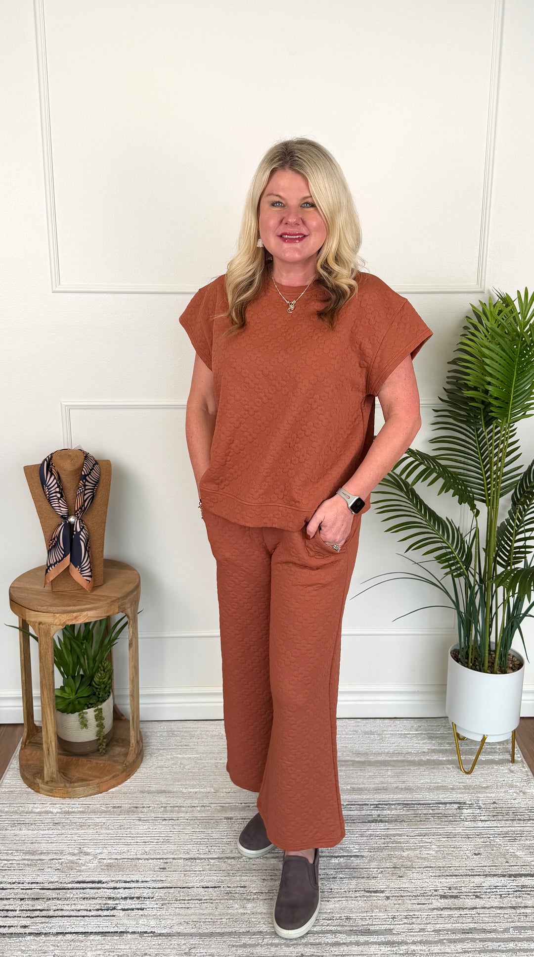 A woman stands indoors modeling a matching rust-orange short-sleeve top and wide-leg pants set. She is posed in front of a white paneled wall with a small wooden side table holding a scarf and a potted palm plant to the right.