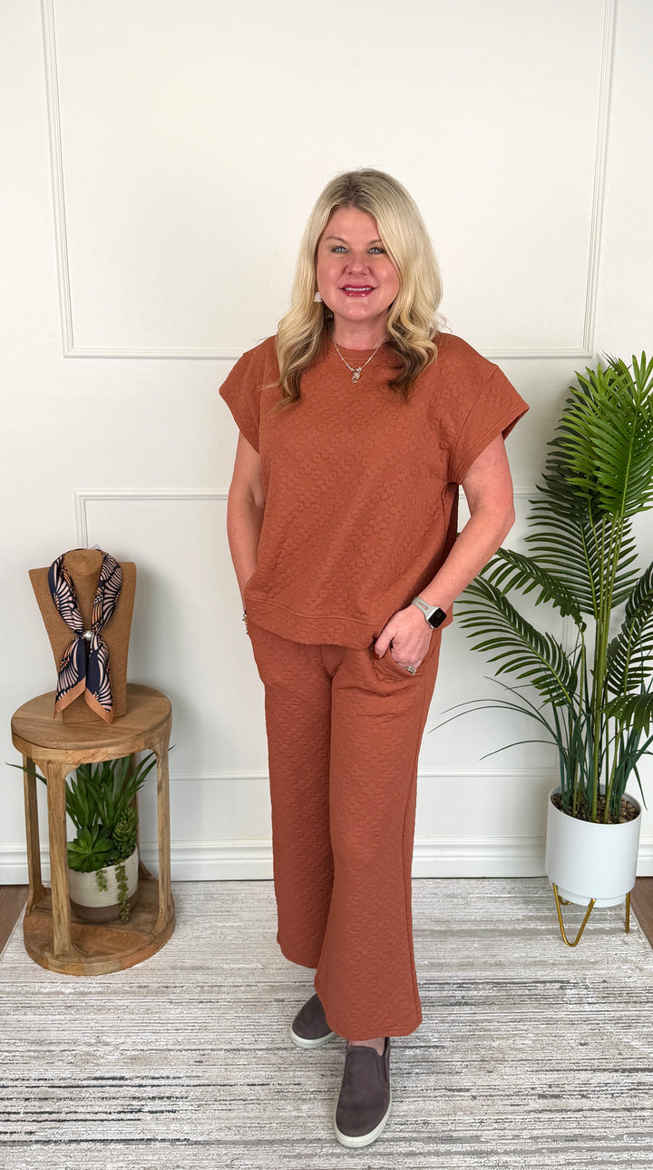 A woman stands indoors modeling a matching rust-orange short-sleeve top and wide-leg pants set. She is posed in front of a white paneled wall with a small wooden side table holding a scarf and a potted palm plant to the right.