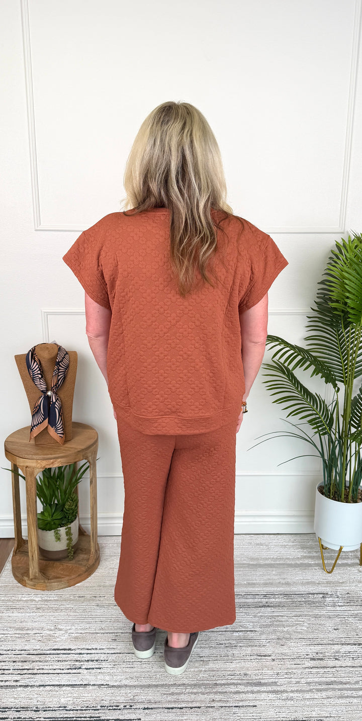 A woman viewed from behind wearing a matching terracotta rust-colored short-sleeve top and wide-leg pants set, standing in a room with a white paneled wall, a wooden side table with a scarf, and a potted palm.