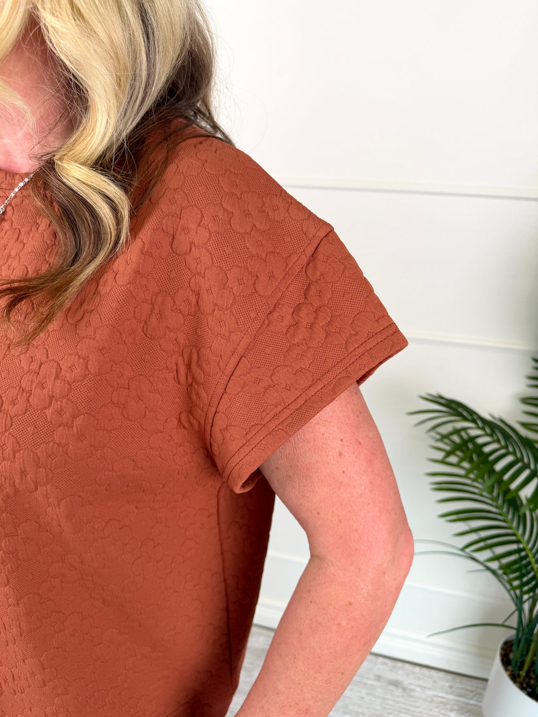 A close-up of a woman wearing a textured terracotta short-sleeve top, showing the fabric's raised pattern and the side of her arm against a white paneled wall with a green plant in the background.