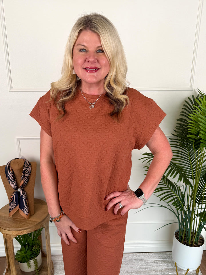 A woman modeling a rust-orange textured short-sleeve top and matching pants set, standing in a styled indoor setting with a white paneled wall, a potted plant, and a small wooden side table.