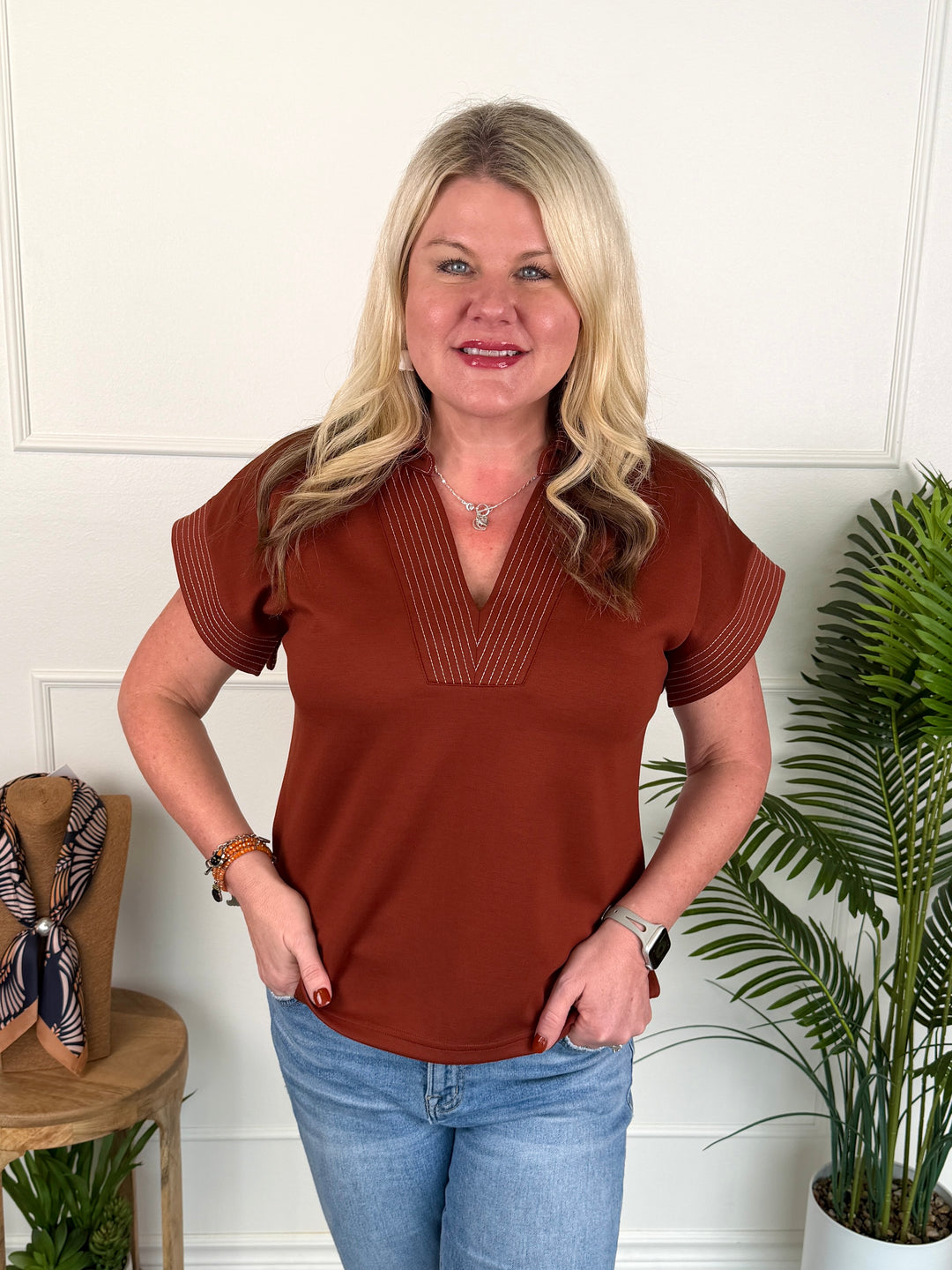 A woman with blonde hair wearing a rust-brown short-sleeve top with pleated detailing on the chest and sleeves, paired with blue jeans, standing in front of a white paneled wall with indoor plants.