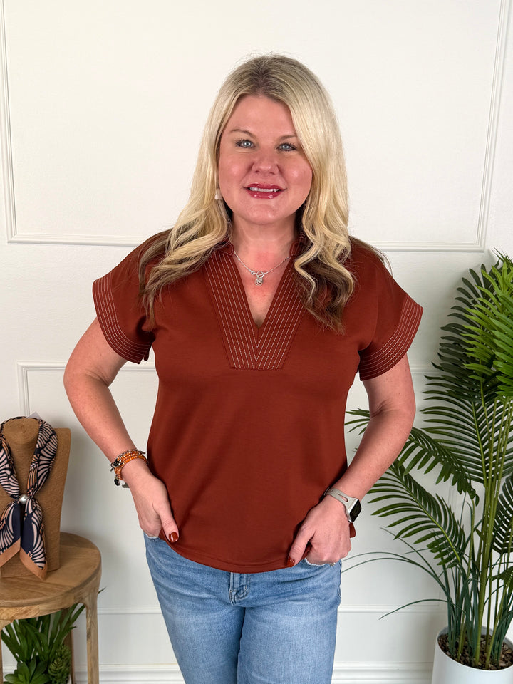 A woman with blonde hair wearing a rust-brown short-sleeve top with pleated detailing on the chest and sleeves, paired with blue jeans, standing in front of a white paneled wall with indoor plants.