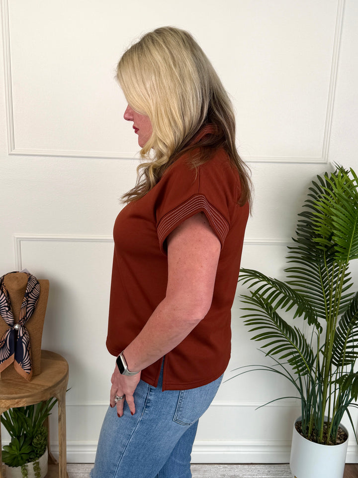 A woman with blonde hair is shown from the side profile wearing a rust-colored short-sleeve knit top and light blue jeans, standing in a room with a white paneled wall, a potted plant, and a small wooden side table.