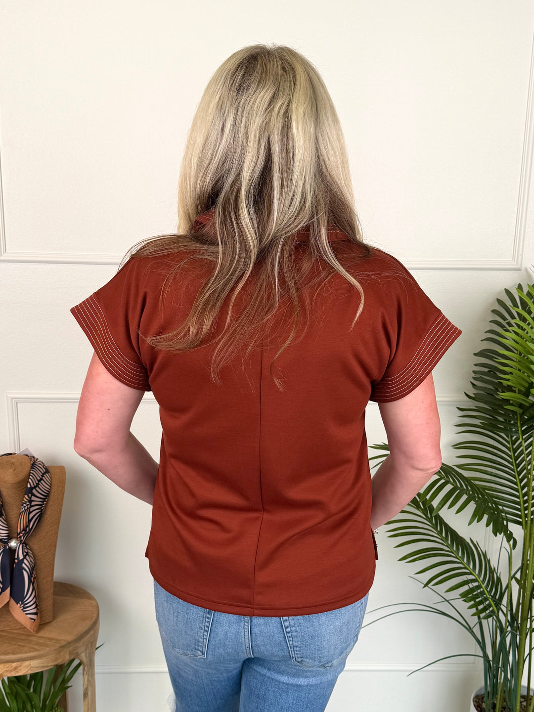A woman viewed from behind wearing a rust-colored short-sleeve top and blue jeans, standing indoors against a white paneled wall with a potted plant to the right.