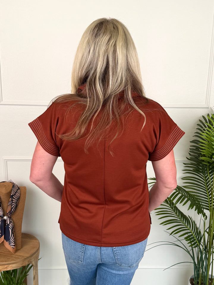 A woman viewed from behind wearing a rust-colored short-sleeve top and blue jeans, standing indoors against a white paneled wall with a potted plant to the right.