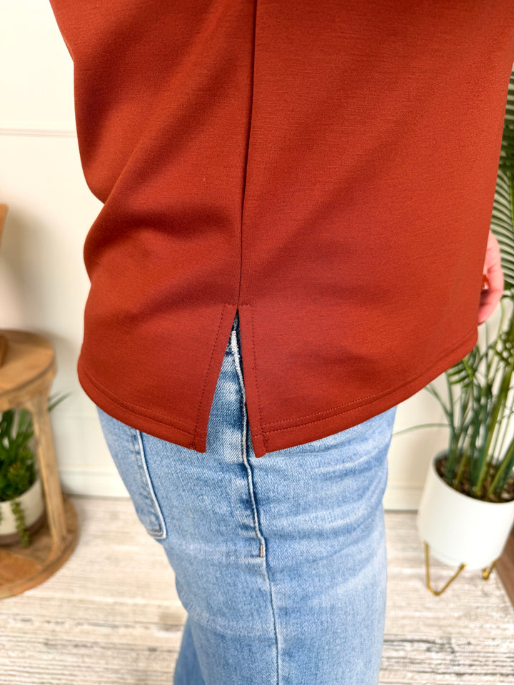 A close-up of a person wearing a rust-orange knit top with a front split V-neck and light-wash blue jeans, standing indoors near potted plants.