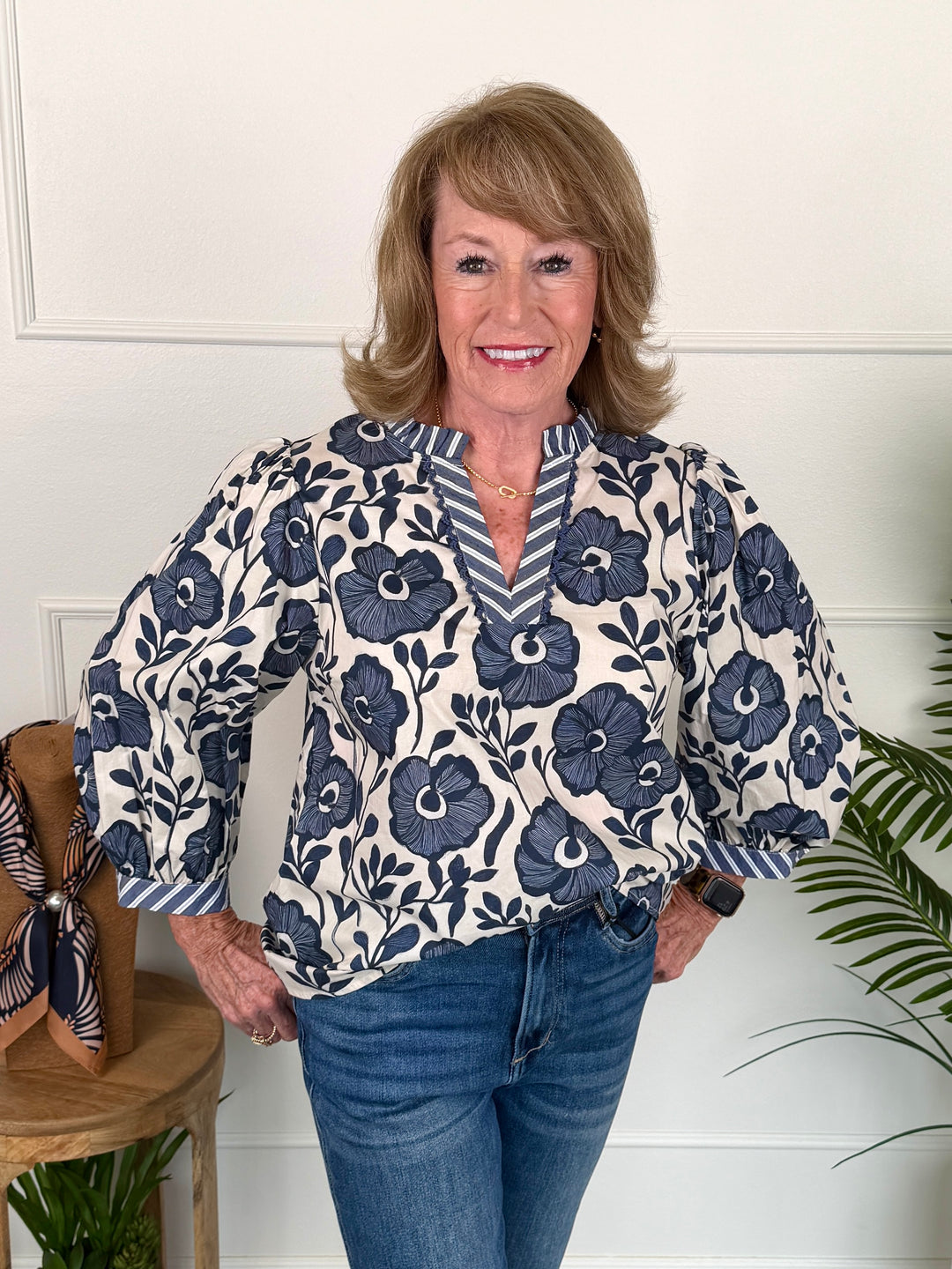 A smiling woman with blonde hair stands indoors wearing a cream blouse with a navy floral print and blue jeans. The blouse has a V-neck with striped trim and elbow-length sleeves.