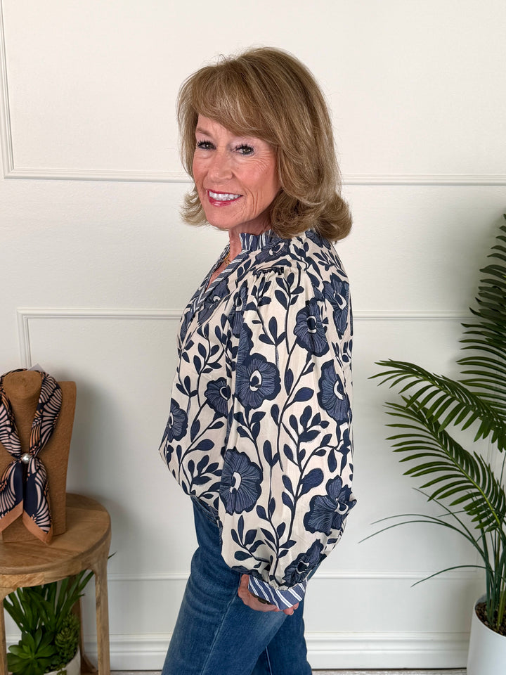 A woman modeling a long-sleeve blouse with a navy blue floral pattern on a cream background, paired with blue jeans, standing in a brightly lit room with white paneled walls and indoor plants.