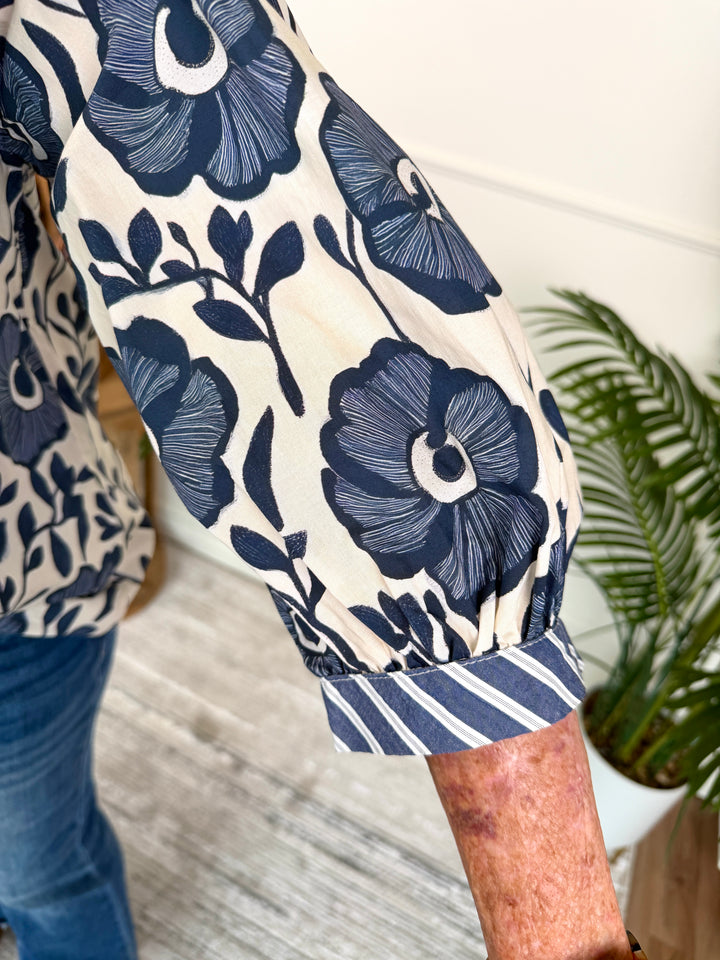 Close-up of a person wearing a cream-colored blouse with a navy blue floral pattern and a striped cuff, paired with blue jeans in an indoor setting.