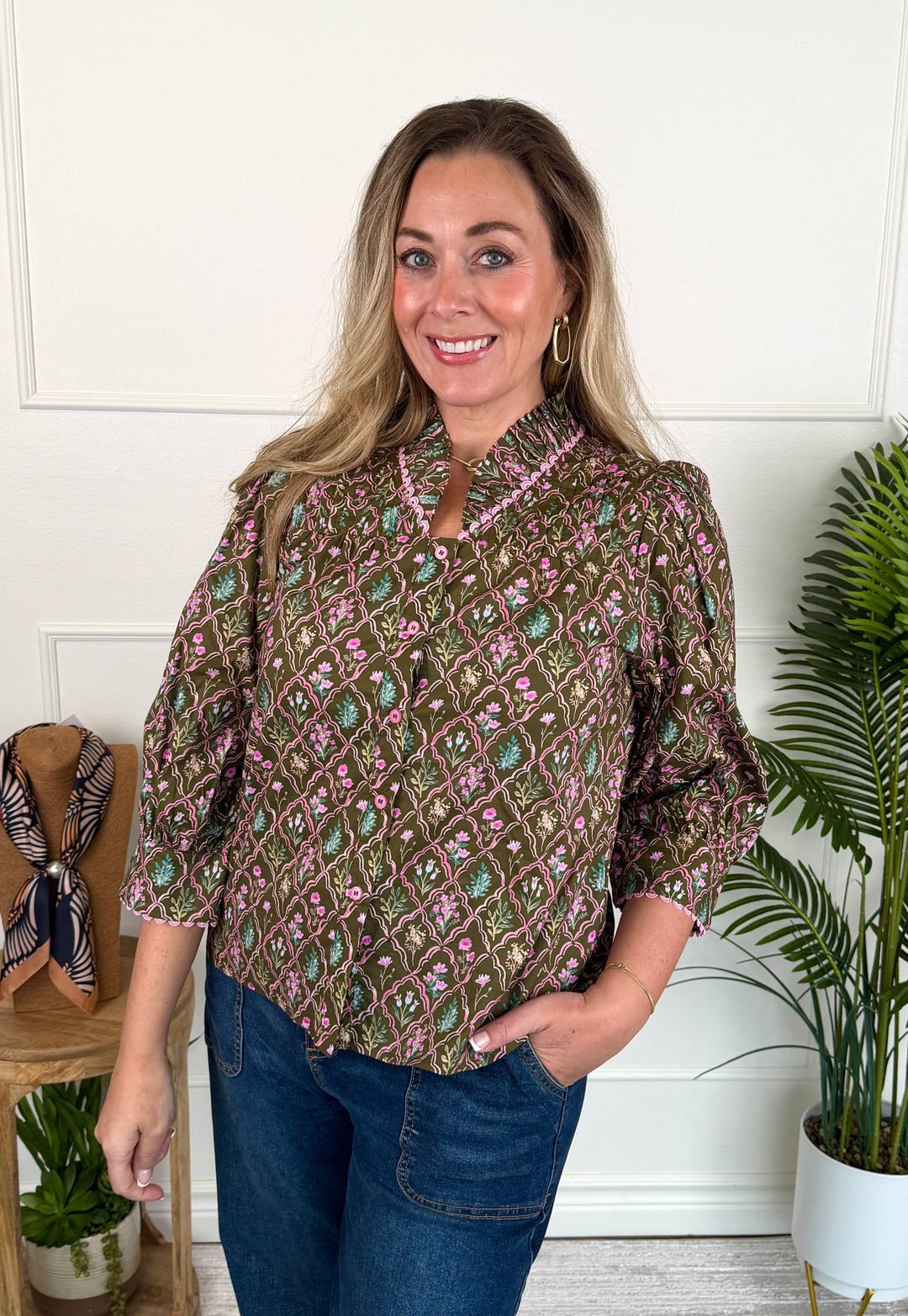 A woman modeling a brown patterned blouse with pink and green floral motifs, paired with blue jeans, standing in a brightly lit room with white paneled walls and potted plants.