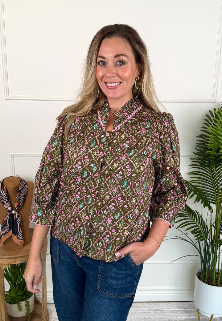 A woman modeling a brown patterned blouse with pink and green floral motifs, paired with blue jeans, standing in a brightly lit room with white paneled walls and potted plants.