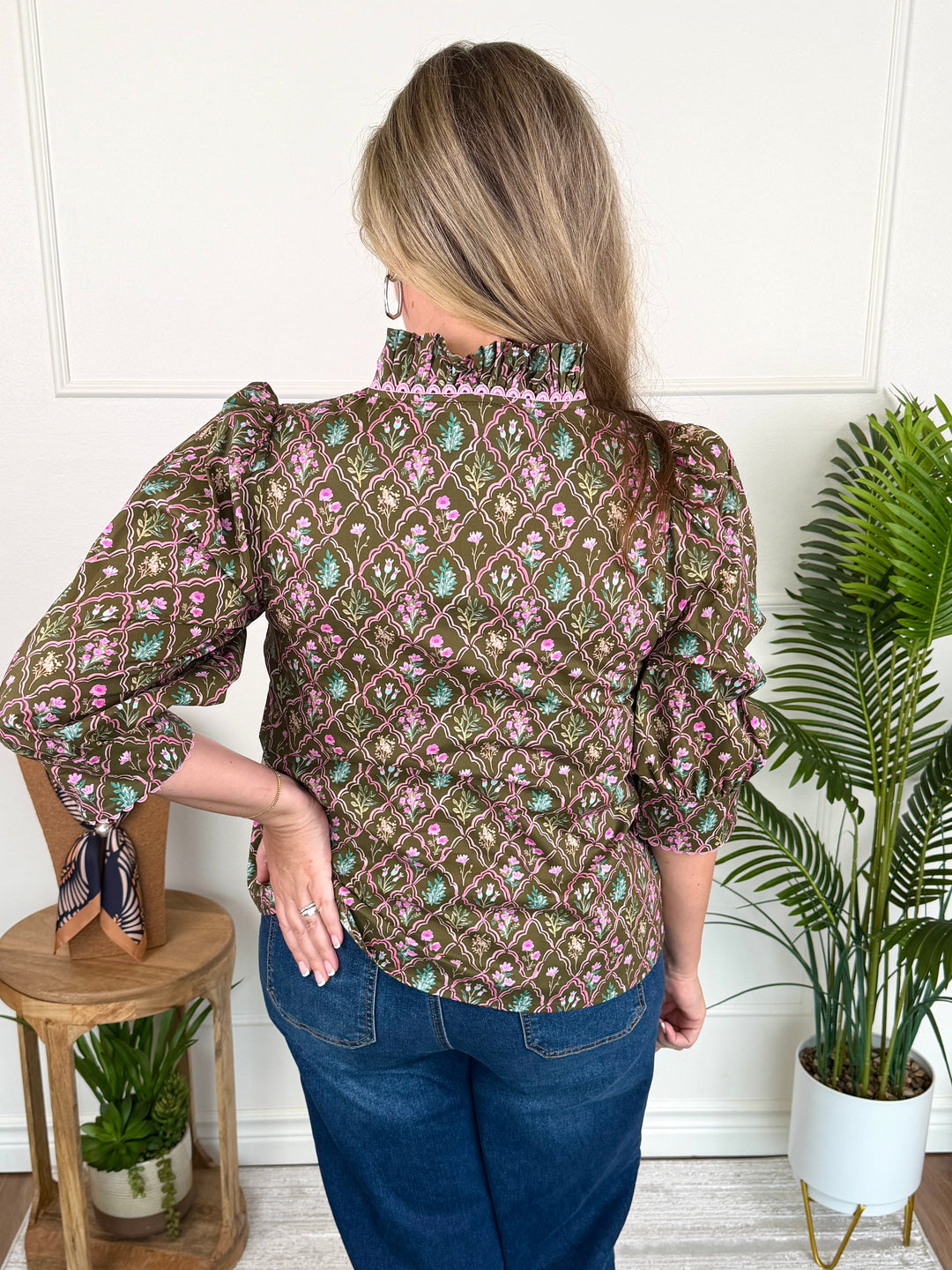 A woman shown from the back wearing a brown patterned blouse with pink and green floral motifs, blue jeans, and a patterned neck scarf, standing in a room with a white wall, a wooden stool, and a potted plant.