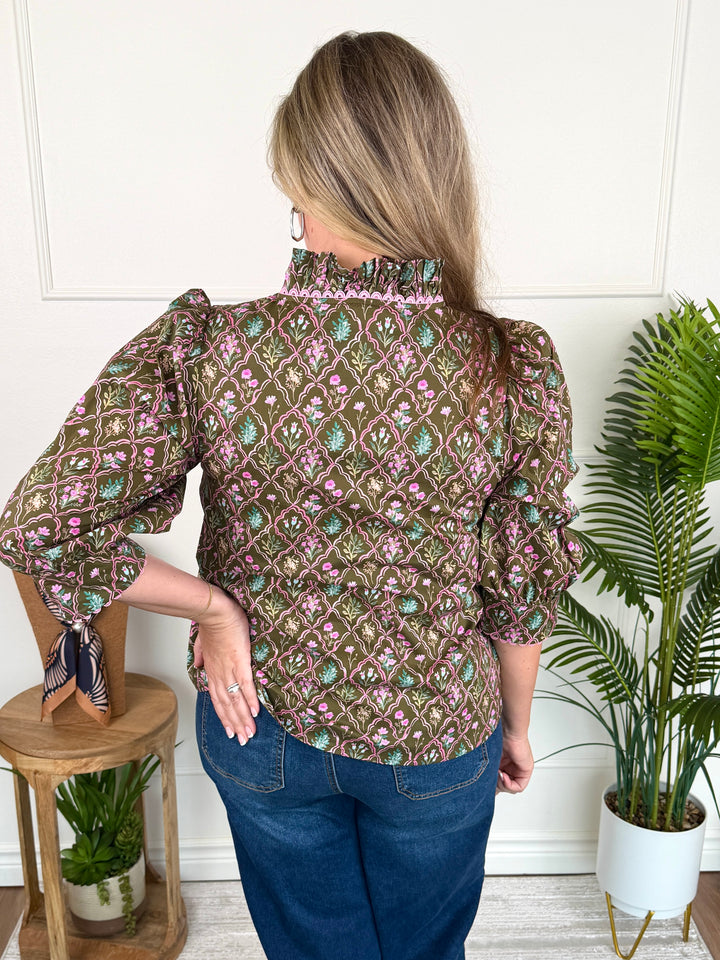 A woman shown from the back wearing a brown patterned blouse with pink and green floral motifs, blue jeans, and a patterned neck scarf, standing in a room with a white wall, a wooden stool, and a potted plant.