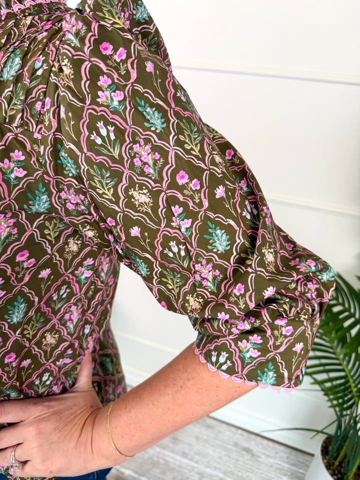 A close-up of a person wearing a brown patterned blouse with pink, green, and white floral and geometric motifs, paired with blue jeans and a gold bracelet, standing indoors near a white paneled wall and a potted plant.