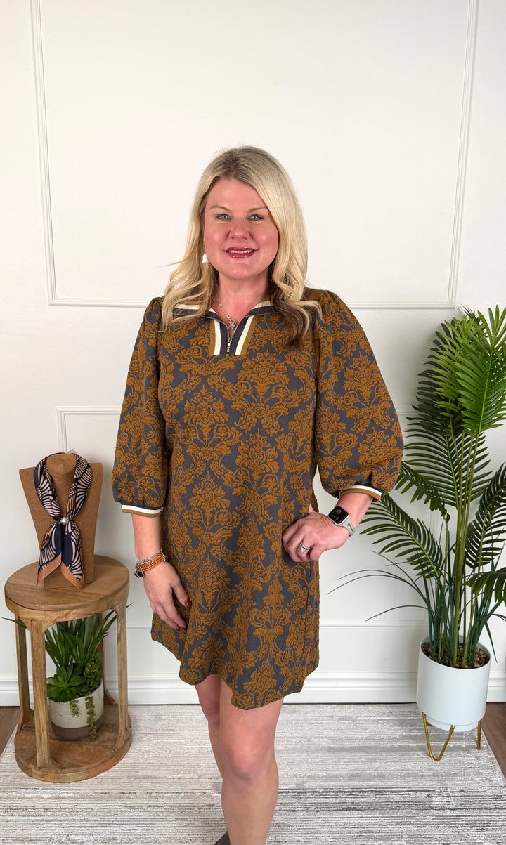 A woman stands indoors wearing a short-sleeve dress with a brown and navy blue floral pattern, a white collar trim, and white cuffs. She is posed in front of a white paneled wall with a small wooden side table on the left and a potted plant on the right.