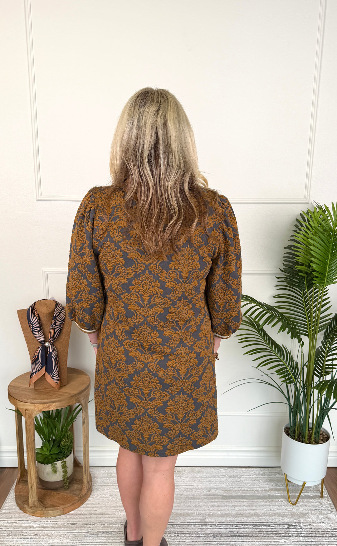 A woman viewed from behind wearing a brown and gold damask-patterned dress, standing in a room with a white paneled wall, a wooden side table with a scarf, and a potted palm plant.