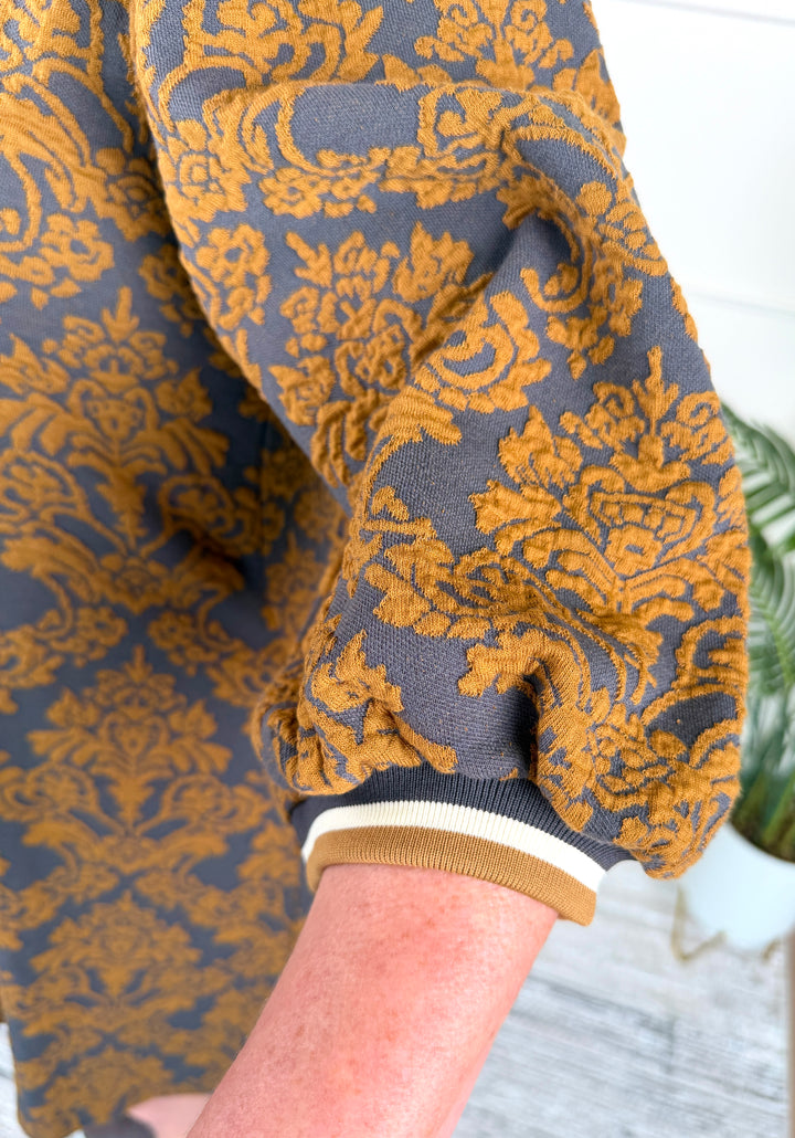 Close-up of a person wearing a gray knit top with a bold mustard yellow damask pattern and a ribbed tan, white, and gray cuff.