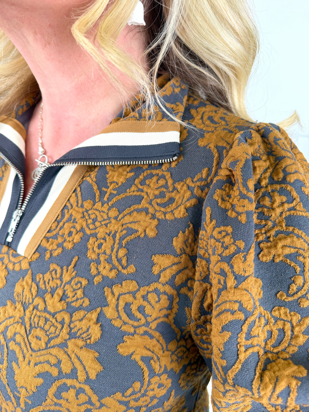 Close-up of a woman wearing a textured jacquard top with a blue-gray base and a golden-yellow damask floral pattern, featuring a mock neck with navy, white, and gold stripes and a silver zipper.
