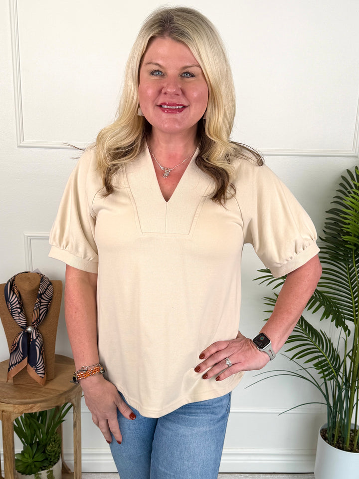 A woman modeling a cream-colored short-sleeve blouse with puff sleeves and a V-neckline, paired with blue jeans, standing in a brightly lit room with indoor plants.
