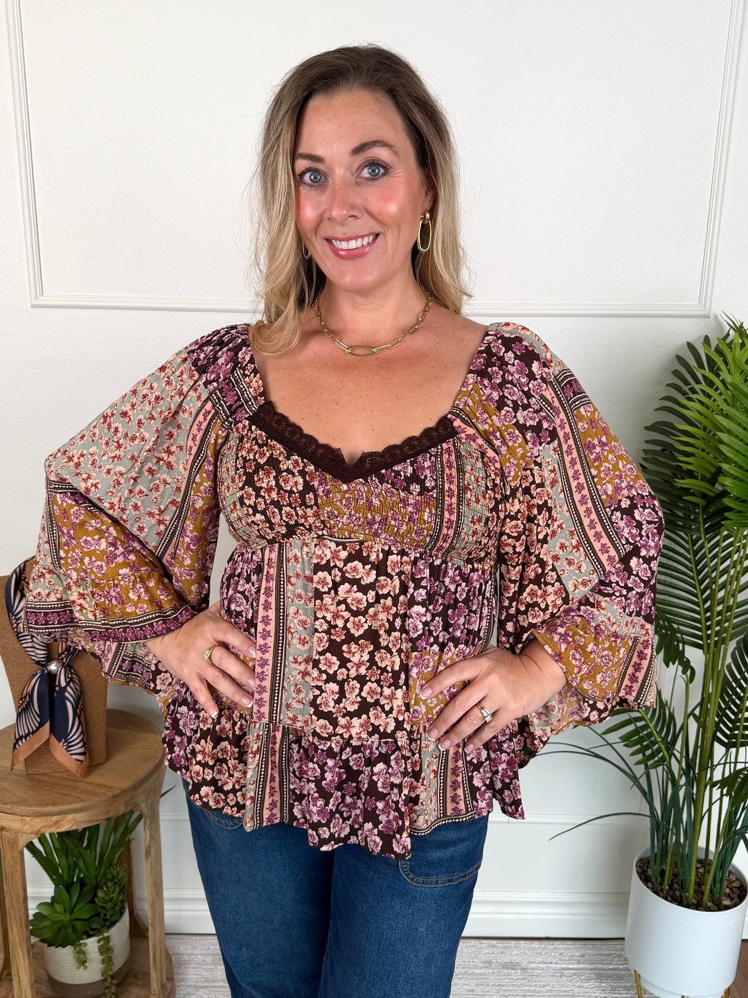 A woman modeling a bohemian-style floral print blouse with wide bell sleeves and a ruffled peplum hem, paired with blue jeans, standing in a brightly lit room with indoor plants.