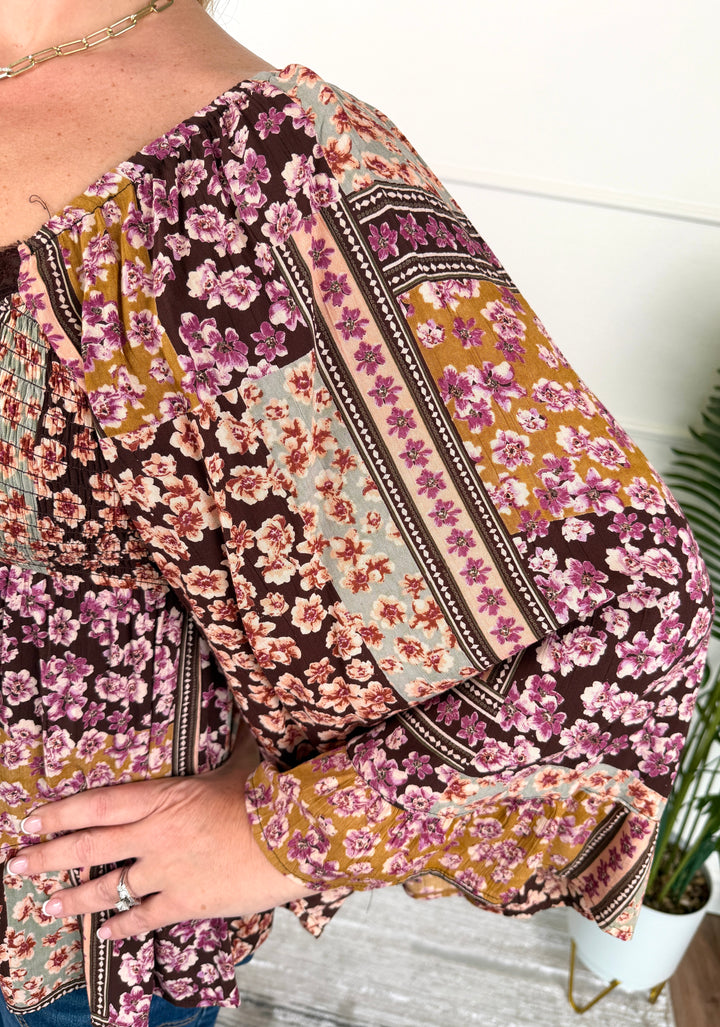A close-up of a woman wearing a long-sleeve blouse with a mixed floral and geometric patchwork print in brown, pink, purple, and mustard tones.